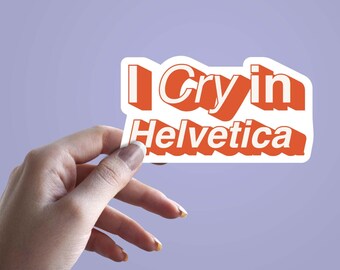 May include: A white sticker with the text "I Cry in Helvetica" in orange, held by a hand against a purple background. The sticker's text is in a bold, sans-serif font.