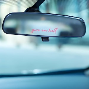 Puede incluir: Un espejo retrovisor de coche con la frase "give em hell" en escritura rosa. El espejo es rectangular con esquinas redondeadas y está montado en el parabrisas. El fondo es una vista borrosa del interior del coche.