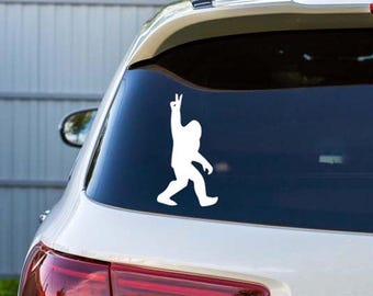 May include: A white vinyl sticker of a Bigfoot figure, giving a peace sign, affixed to the rear window of a white car. The silhouette of the creature is in a walking pose, with one arm raised in a peace gesture.