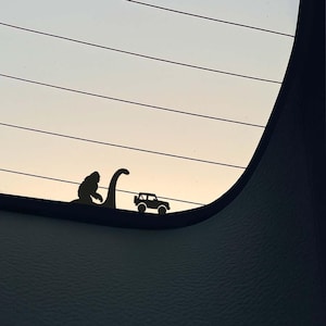 May include: Black vinyl car decal featuring silhouettes of a Bigfoot, a Loch Ness Monster, and a Jeep. The decal is placed on a car window with a sunset background. The design is simple and humorous.