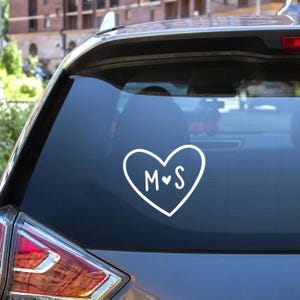 Puede incluir: Calcomanía blanca en forma de corazón con las letras "M" y "S" en su interior. La calcomanía está adherida a la ventana trasera de un vehículo. El corazón está delineado con un diseño simple y las letras son manuscritas.