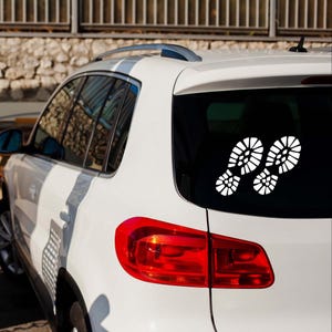 Puede incluir: Calcomanía blanca de dos huellas de pies en la ventana trasera de un SUV blanco. Las huellas tienen patrones de banda de rodadura detallados, lo que sugiere un tema de senderismo o al aire libre. El coche está aparcado fuera.