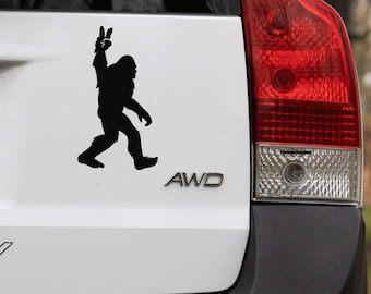 May include: Black silhouette of Bigfoot giving a peace sign, applied to a white vehicle. The image shows the creature walking with one arm raised, displaying the peace symbol. The car's taillight and the letters "AWD" are also visible.