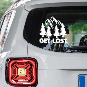 May include: White vinyl decal featuring a mountain range and pine trees. The text "GET LOST" is below the graphic. The decal is applied to the rear window of a white vehicle.