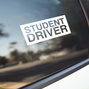 May include: A white rectangular sticker with the words "STUDENT DRIVER" in bold, black capital letters. The sticker is affixed to a car window, with a blurred outdoor scene visible through the glass.