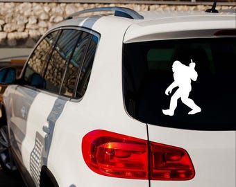 May include: A white vinyl decal of Bigfoot giving the middle finger is affixed to the rear window of a white SUV. The decal is a silhouette of the legendary creature, with its arm raised in a gesture of defiance.