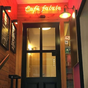May include: Entrance to a cafe with a black door and glass panels. The neon sign above the door reads "Café falala". The walls are wood-paneled and painted red. Two sconces provide additional lighting.