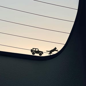 May include: A black vinyl decal of a small SUV and a cat jumping. The decal is applied to a car window, with the sky visible in the background. The cat appears to be chasing the SUV.