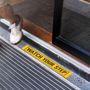 Peut inclure: Un panneau d'avertissement rectangulaire jaune avec le texte "WATCH YOUR STEP" en lettres majuscules blanches. Le panneau est placé sur une surface rayée grise et noire près d'une porte. On voit un pied et une chaussure.