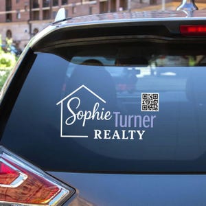 May include: White vinyl decal on a car window featuring the name "Sophie Turner" in a script font, with a house outline and the word "REALTY" below. A QR code is on the right. The decal is on a car's rear window.