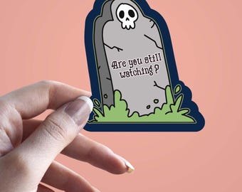 Peut inclure: Un autocollant représentant une pierre tombale de dessin animé avec un crâne et le texte "Are you still watching ?" La pierre tombale est grise avec un bord bleu foncé et de l'herbe verte. L'autocollant est tenu par une main sur un fond rose.