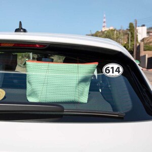 Op de afbeelding: Een lichtgroene, getextureerde make-uptas met een oranje rits hangt in een auto. Een witte ovale sticker met het nummer "614" staat op de autoruit. De achtergrond toont een verre toren en bomen.