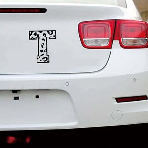 May include: A black and white letter "T" decal with a cow print design is affixed to the rear of a white car. The decal is centered on the trunk, with the car's taillights visible on the right side.
