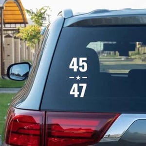 以下が含まれることがあります： 車の窓に貼られた白いビニールステッカー。数字の「45」が星とダッシュのデザインの上にあり、その下に数字の「47」があります。車は濃い色で、背景には住宅街が写っています。