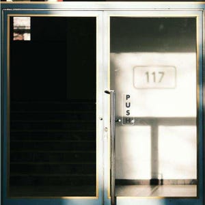 May include: A glass door with a silver handle and a "PUSH" sign. The number "117" is displayed on the glass. The door frame is blue with gold trim. Stairs are visible through the left door.