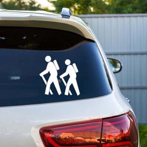 Puede incluir: Adhesivo de vinilo blanco de dos excursionistas con mochilas y bastones de senderismo, aplicado en la ventana trasera de un coche. Las figuras son estilizadas. El coche es blanco.