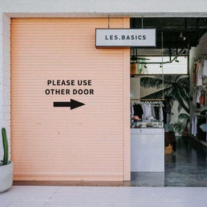 May include: Exterior shot of a clothing store with a peach-colored, closed, slatted door. The sign above the door reads "LES.BASICS." Text on the door reads "PLEASE USE OTHER DOOR" with an arrow pointing right. A cactus in a white pot is on the left.