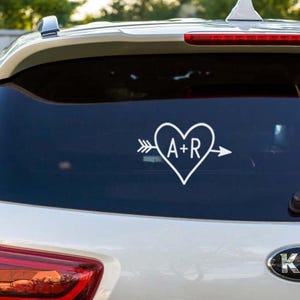 May include: White vinyl car decal featuring a heart with the letters "A+R" inside, pierced by an arrow. The decal is displayed on a car's rear window. The design is simple and elegant, suitable for personalizing a vehicle.