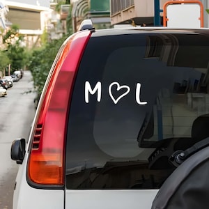 Może przedstawiać: Biała naklejka na samochód z literami "M", symbolem serca i literą "L". Naklejka jest naklejona na tylnej szybie białego samochodu. Tylne światło samochodu jest czerwone.
