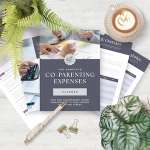 Puede incluir: Un planificador titulado "The Complete Co-Parenting Expenses" con una cubierta gris oscuro, rodeado de otros documentos, un bolígrafo y una taza de café. La portada del planificador incluye la palabra "Planner" y el texto "Fair and Transparent Money Management to Ease Shared Parenting Stress."