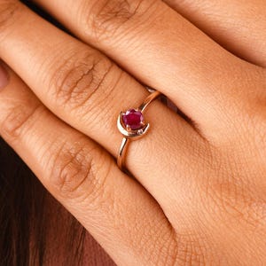 May include: A gold ring with a small, round, red gemstone. The ring is on a person's finger, with the hand resting near the chest. The person is wearing a pink shirt.