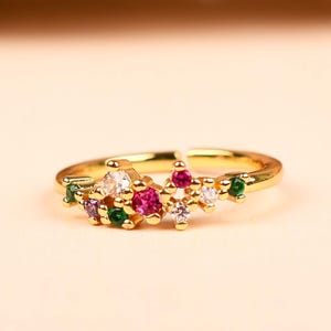 May include: A gold ring with a cluster of colourful gemstones. The ring has a slender band and is decorated with pink, green, white, and purple stones, forming a floral or celestial pattern. The gemstones are closely set together.
