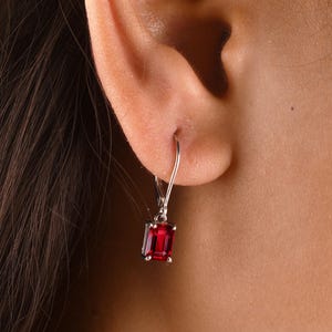 May include: A silver-toned leverback earring featuring a rectangular, emerald-cut deep red gemstone. The earring is suspended from a metal hook that secures through the earlobe, showcasing a faceted stone with a polished metallic setting.