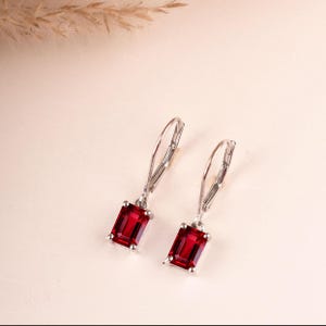May include: A pair of silver leverback earrings, each featuring a single rectangular emerald-cut deep red gemstone suspended from a polished metal hook. The earrings are designed with a simple prong setting that secures the vibrant red stones.