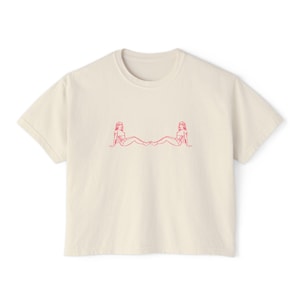 May include: Cream-colored t-shirt featuring a red line drawing of two figures facing each other. The figures are in a seated position. The t-shirt has a crew neck and short sleeves.