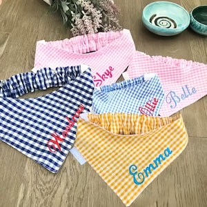 May include: A collection of pet bandanas in various gingham patterns, including pink, blue, and yellow. Each bandana is personalized with a name embroidered in a contrasting color. The bandanas are arranged on a wooden surface.