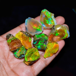 May include: A selection of rough opal gemstones, displaying a spectrum of colours. The opals exhibit yellow, orange, green, and blue tones, with a play of colour effect. The gemstones are held in a hand against a dark backdrop.
