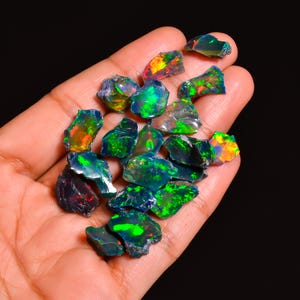 Könnte beinhalten: Eine Sammlung roher, ungeschliffener Opale in verschiedenen Formen und Größen. Die Opale zeigen ein lebendiges Farbspiel mit grünen, blauen, orangen und gelben Blitzen. Die Edelsteine werden in einer Hand vor einem schwarzen Hintergrund gehalten.