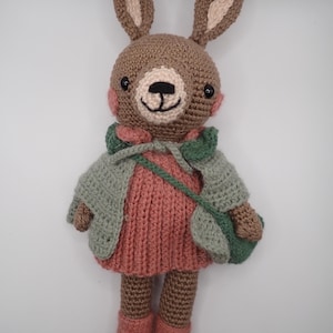 Könnte beinhalten: Handgemachte Häkelkaninchenpuppe mit rosa Kleid, grüner Jacke und grüner Handtasche. Das Kaninchen hat braunes Fell, rosa Wangen und lange Ohren. Die Puppe ist etwa 25 cm groß.