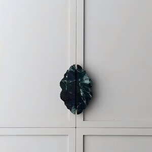 May include: A pair of dark green marble cabinet pulls with a scalloped, leaf-like edge. The two pieces are mounted vertically on a light gray cabinet door, meeting in the center to form a single, symmetrical, oval-shaped decorative handle.