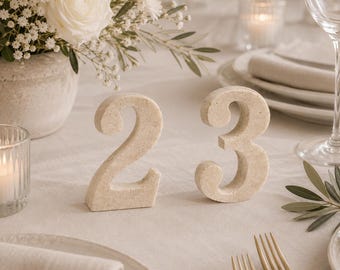 May include: A pair of freestanding table numbers, specifically the digits two and three, crafted from a light beige, textured stone-like material. The numbers feature a smooth, rounded font and are positioned upright on a flat surface.