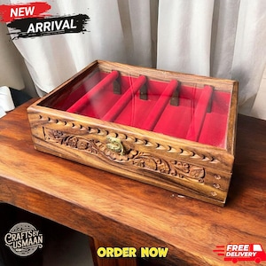 May include: A rectangular wooden jewelry box with a clear lid and red velvet interior. The box features intricate carved floral designs and a brass clasp. The words "NEW ARRIVAL" and "ORDER NOW" are visible.