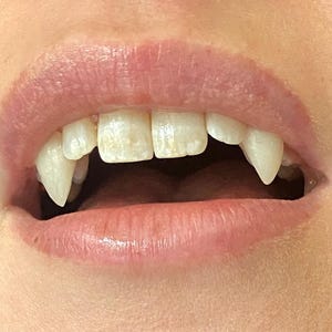 May include: Close-up of a mouth with pale pink lips and teeth. Two sharp, pointed vampire fangs are visible, along with other teeth. The image is well-lit, highlighting the details of the teeth and lips.