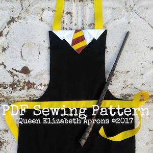 May include: Black apron with yellow straps and a red and yellow tie design. The apron has a white collar and a small label that says "Queen Elizabeth Aprons ©2017". A wooden wand is lying on top of the apron.