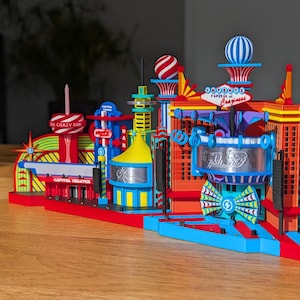 Può includere: Un colorato modello in miniatura di una città con edifici, insegne ed elementi decorativi. Il modello presenta una base rossa, con edifici blu, gialli e rossi. Il testo "The Crazy Inn" e "Capital of Craziness" sono visibili.