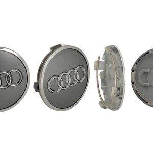Könnte beinhalten: Vier Audi Radnabenabdeckungen. Die Kappen sind dunkelgrau mit einem silbernen Audi-Logo. Eine Kappe wird von der Seite gezeigt und zeigt die Rückseite und den Befestigungsmechanismus. Die Kappen sind rund und haben ein glänzendes Finish.