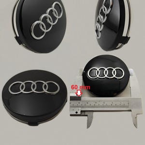 Può includere: Quattro coprimozzi Audi neri con logo Audi argentato. I coprimozzi sono mostrati da diverse angolazioni, uno dei quali viene misurato a 60 mm con un calibro. I coprimozzi sono lucidi.