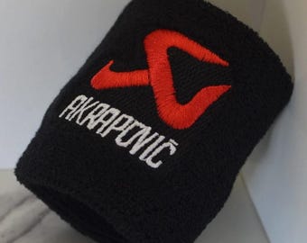 Peut inclure: Poignet de sport noir avec le texte "AKRAPOVIČ" brodé en blanc et un logo rouge. Le bracelet est fait d'un matériau doux et absorbant, probablement en éponge, et est conçu pour absorber la sueur pendant l'activité physique.