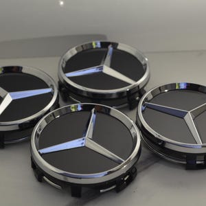 Puede incluir: Cuatro tapacubos negros y plateados con el logotipo de Mercedes-Benz. Cada tapacubos presenta un centro negro con un emblema de estrella de tres puntas plateada, rodeado por un borde plateado. Los tapacubos se muestran sobre una superficie clara.