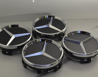 Peut inclure: Quatre enjoliveurs noirs et argentés avec le logo Mercedes-Benz. Chaque enjoliveur présente un centre noir avec un emblème en étoile à trois branches argenté, entouré d'une jante argentée. Les enjoliveurs sont présentés sur une surface claire.