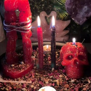 May include: A collection of lit candles, including a red skull-shaped candle, a large red candle depicting two figures embracing, and several tall, slender candles. The candles are surrounded by dried flower petals and small candles, creating a mystical ambiance.