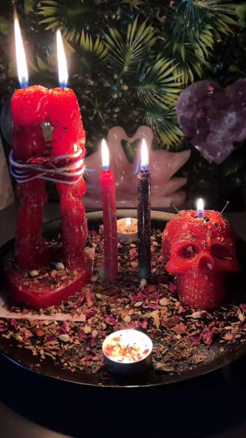 May include: A collection of lit candles, including a red skull-shaped candle, a large red candle depicting two figures embracing, and several tall, slender candles. The candles are surrounded by dried flower petals and small candles, creating a mystical ambiance.