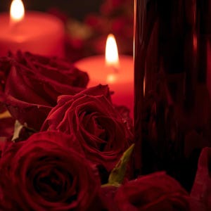May include: A romantic still life featuring red roses, lit candles, and a dark glass bottle. The roses are in full bloom, with soft petals. The candles emit a warm glow, creating a cozy atmosphere. The bottle is partially visible, adding depth to the composition.