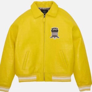 K&ouml;nnte beinhalten: Eine leuchtend gelbe Jacke mit strukturierter Krokodilpr&auml;gung, ausgestattet mit einem durchgehenden Frontrei&szlig;verschluss, einem klassischen Kragen und zwei Seitentaschen. Auf der Brust befindet sich ein Avirex-Wappen, &Auml;rmelb&uuml;ndchen und Saum sind wei&szlig; gestreift.