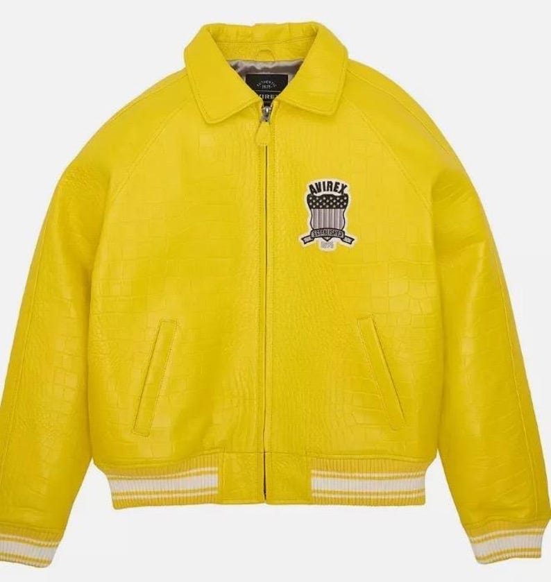 K&ouml;nnte beinhalten: Eine leuchtend gelbe Jacke mit strukturierter Krokodilpr&auml;gung, ausgestattet mit einem durchgehenden Frontrei&szlig;verschluss, einem klassischen Kragen und zwei Seitentaschen. Auf der Brust befindet sich ein Avirex-Wappen, &Auml;rmelb&uuml;ndchen und Saum sind wei&szlig; gestreift.