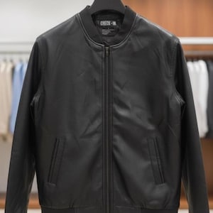 May include: Black bomber jacket with a zip-up front, ribbed collar, cuffs, and hem. The jacket has two front pockets and the text "CHECK-IN" on the collar. The jacket is hanging on a metal hanger.